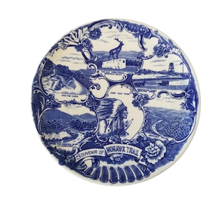 May include: A white ceramic souvenir plate with a blue floral border and illustrations of the Mohawk Trail. The plate features images of a deer, a Native American chief, and scenic views. The text "Souvenir of Mohawk Trail" is displayed.
