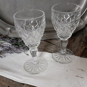 May include: Two clear crystal wine glasses with diamond-cut patterns. The glasses have a stemmed base and a round foot. The background includes a silver tray, lavender sprigs, and a white cloth on a wooden surface.