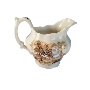 May include: Cream-colored ceramic pitcher with a landscape design. The pitcher features a curved handle and a spout. The landscape design includes trees, a river, and a cloudy sky. The words "The Peak Horse" are printed on the bottom.