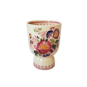 May include: A white ceramic egg cup with a floral design. The cup has a pink, yellow, blue, and green floral pattern. The cup has a pedestal base.