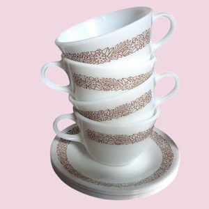May include: A stack of four white teacups and saucers with a brown floral design. The cups are stacked on top of each other, with the top cup tilted slightly. The saucers are stacked at the bottom. The background is a light pink.