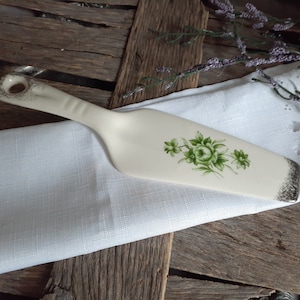 May include: Off-white ceramic cake server with a green floral design. The handle has a hole for hanging and a silver-colored trim. The server rests on a white linen napkin, with a rustic wooden background.
