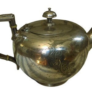 May include: A silver-colored teapot with a rounded body, a curved spout, and a square handle. The lid is topped with a small knob. The teapot has decorative etched patterns on its surface, suggesting an antique design.