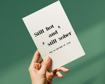 Puede incluir: Una tarjeta blanca con el texto "Still hot and still sober" y "Im so proud of you" en negro. La tarjeta se sostiene en una mano sobre un fondo verde.