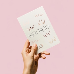 Puede incluir: Tarjeta de felicitación crema con la frase "YOU'RE THE TITS" en letras negras. La tarjeta presenta ilustraciones de pechos en varios tonos de piel. El fondo es de color rosa claro.