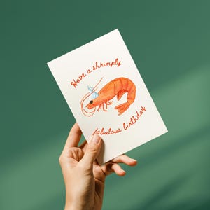 K&ouml;nnte beinhalten: Eine wei&szlig;e rechteckige Gru&szlig;karte mit einer handgezeichneten orangefarbenen Garnele, die einen kleinen blau-wei&szlig; gestreiften Partyhut tr&auml;gt. Der in roter Schreibschrift gedruckte Text lautet, Have a shrimply fabulous birthday, um die Garnele herum.