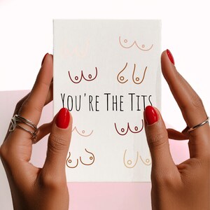 Puede incluir: Una impresi&oacute;n en lienzo blanco con dibujos lineales de senos en varios tonos de piel. El texto "YOU'RE THE TITS" est&aacute; impreso en negro. La impresi&oacute;n est&aacute; sostenida por manos con esmalte de u&ntilde;as rojo y anillos plateados.