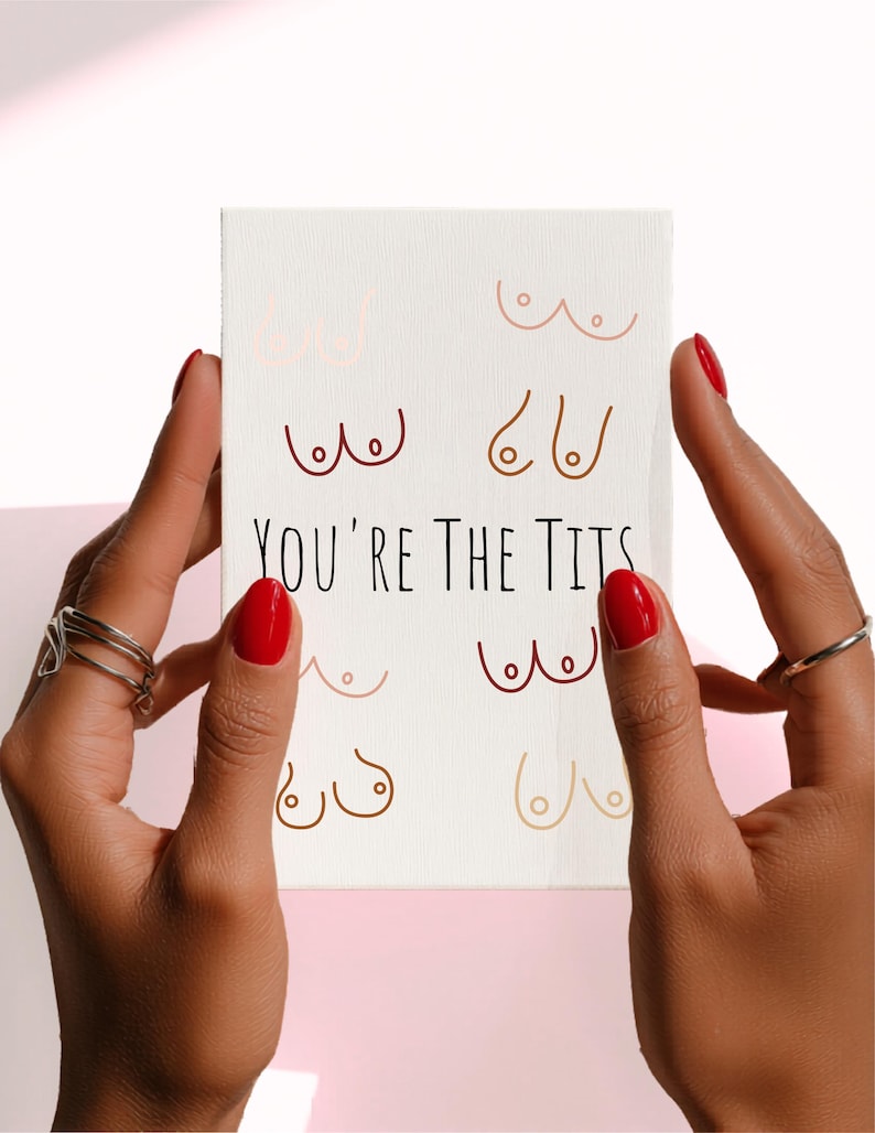 Puede incluir: Una impresi&oacute;n en lienzo blanco con dibujos lineales de senos en varios tonos de piel. El texto "YOU'RE THE TITS" est&aacute; impreso en negro. La impresi&oacute;n est&aacute; sostenida por manos con esmalte de u&ntilde;as rojo y anillos plateados.