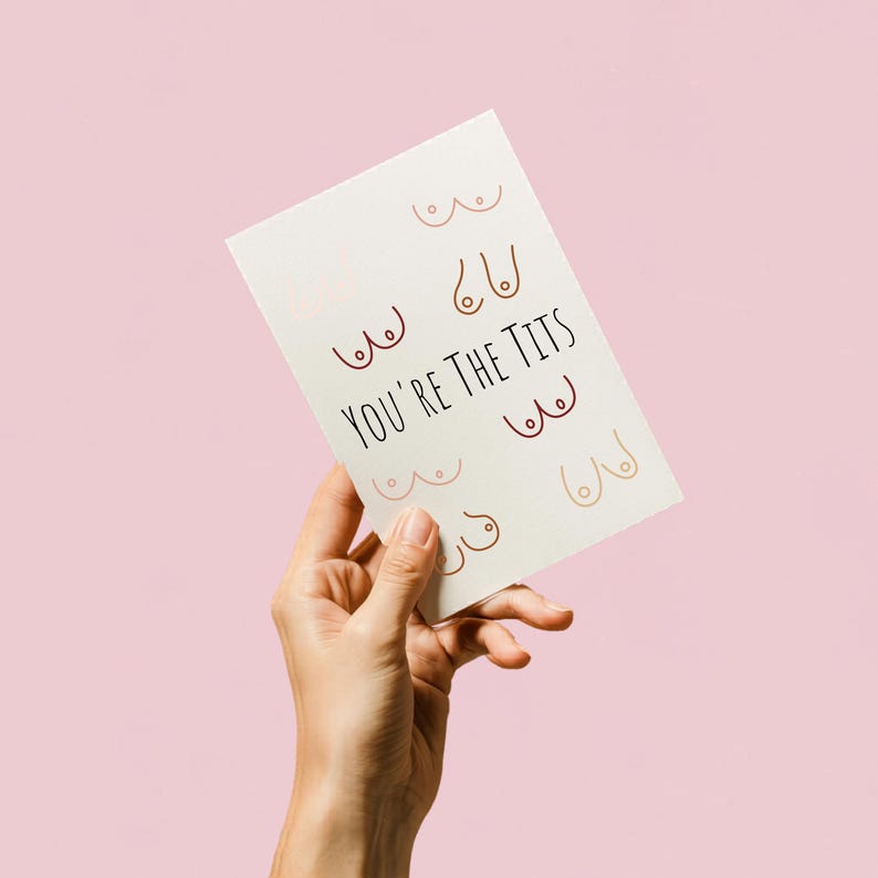 May include: A greeting card with the text "YOU'RE THE TITS" in black lettering. The card features several illustrations of breasts in various skin tones and shapes. The background is a solid pink color.
