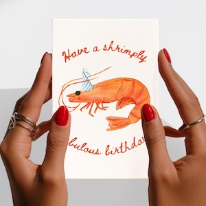 K&ouml;nnte beinhalten: Eine rechteckige Gru&szlig;karte mit wei&szlig;em Hintergrund und einer Aquarellillustration einer orangefarbenen Garnele, die einen blau-wei&szlig; gestreiften Partyhut tr&auml;gt. Der Text lautet Have a shrimply fabulous birthday in orangefarbener Schreibschrift.