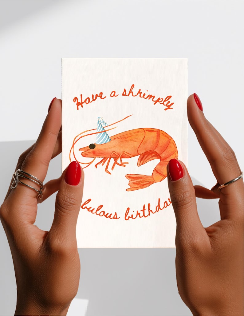 K&ouml;nnte beinhalten: Eine rechteckige Gru&szlig;karte mit wei&szlig;em Hintergrund und einer Aquarellillustration einer orangefarbenen Garnele, die einen blau-wei&szlig; gestreiften Partyhut tr&auml;gt. Der Text lautet Have a shrimply fabulous birthday in orangefarbener Schreibschrift.