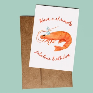 K&ouml;nnte beinhalten: Eine wei&szlig;e Gru&szlig;karte mit einer orangefarbenen Garnelenillustration, die einen blau-wei&szlig; gestreiften Partyhut tr&auml;gt. Der Text lautet Have a shrimply fabulous birthday in orangefarbener Schreibschrift. Die Karte liegt auf einem braunen Kraftpapierumschlag.