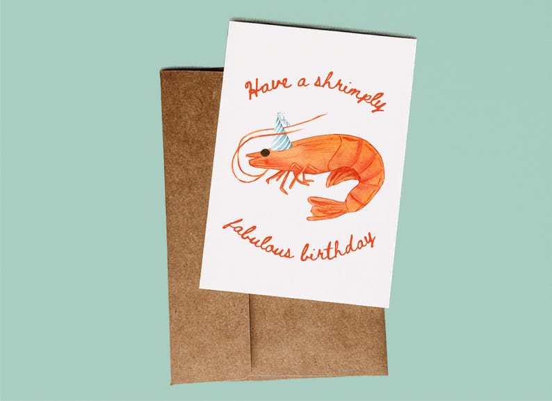 K&ouml;nnte beinhalten: Eine wei&szlig;e Gru&szlig;karte mit einer orangefarbenen Garnelenillustration, die einen blau-wei&szlig; gestreiften Partyhut tr&auml;gt. Der Text lautet Have a shrimply fabulous birthday in orangefarbener Schreibschrift. Die Karte liegt auf einem braunen Kraftpapierumschlag.