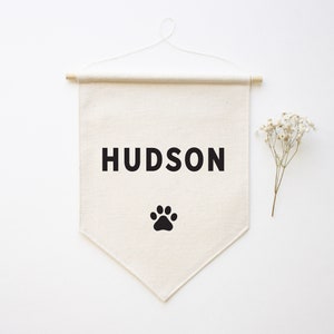 Puede incluir: Una pancarta de lona blanca con el nombre "HUDSON" impreso en letras negras y una huella de pata negra debajo. La pancarta está colgada de una varilla de madera.