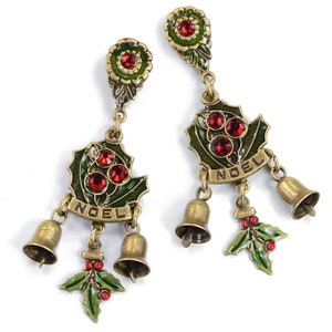 May include: A pair of dangle earrings with a green holly leaf design, red crystals, and gold bells. The earrings have the word "NOEL" written on them.