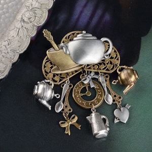 Puede incluir: Una tetera, una taza de té y otros encantos con temática de té de plata y oro cuelgan de un diseño de filigrana. Los encantos incluyen una esfera de reloj, cucharas, un corazón y una tetera más pequeña.