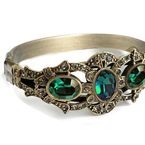 May include: A gold-toned bangle bracelet with an ornate design featuring three emerald green gemstones.