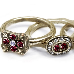May include: Set of six silver rings with red gemstone accents. The rings feature various styles, including a simple band with a single stone, a ring with a floral design, and a ring with a cluster of stones.