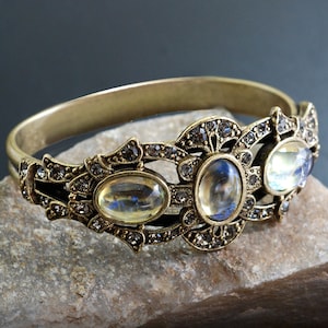 May include: A gold-toned bangle bracelet with a decorative design featuring three oval-shaped clear stones and smaller rhinestones.