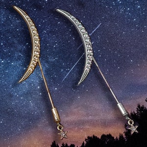 Puede incluir: Dos broches de media luna, uno dorado y otro plateado, cada uno adornado con pedrería transparente. De cada luna se extiende un fino alfiler, que termina en un pequeño dije de estrella. El fondo presenta un cielo estrellado.