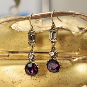 May include: A pair of gold tone dangle earrings with purple, clear, and gray faceted stones. The earrings have a teardrop shape and are accented with a small, round stone.