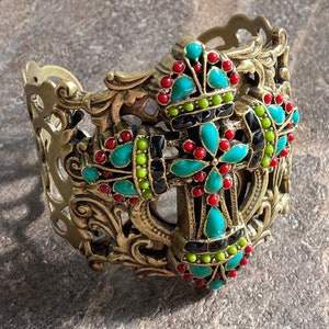May include: A gold-tone metal cuff bracelet with an ornate design featuring a turquoise, red, and black cross. The cross is embellished with colorful beads.