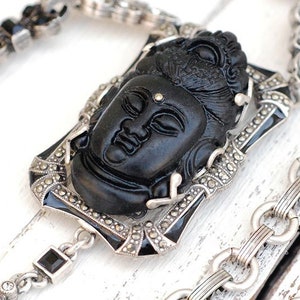 May include: A silver chain necklace with a black carved Buddha pendant. The pendant is set in a silver frame with black enamel accents. The chain is made of small silver links.