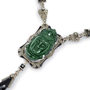 May include: A silver necklace with a green jade pendant carved with a face. The pendant is surrounded by black and white stones. The necklace is on a white background.