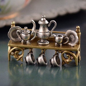 Puede incluir: Un broche de plata y oro con un juego de té con tetera, jarra de crema, azucarero y cuatro tazas colgando de un estante. El broche está decorado con volutas ornamentadas y es perfecto para los amantes del té.