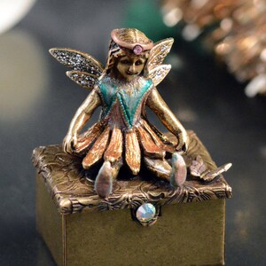 May include: A gold-toned trinket box with a fairy figurine on top. The fairy is wearing a green and orange dress with a pink crown and has wings with gold and green accents. The box has a decorative design and a clear rhinestone in the center.