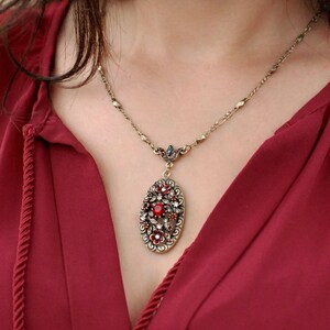 May include: A gold chain necklace with a red and clear crystal pendant. The pendant is oval shaped and features a floral design.