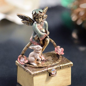 May include: A small, rectangular, gold-colored trinket box with a fairy and rabbit figurine on top. The fairy is wearing a green and pink outfit, and the rabbit is white. The box has a jeweled clasp and floral accents.