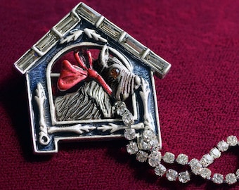 Peut inclure: Une broche argentée en forme de niche à chien avec un chien à l'intérieur. Le chien porte un nœud rouge et un collier en strass. La niche a un toit bleu et des bordures blanches. Une chaîne en strass est attachée à la niche.