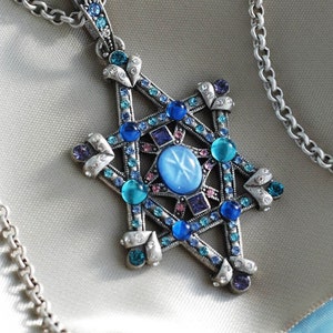 Może przedstawiać: Srebrny naszyjnik z zawieszką w kształcie gwiazdy. Zawieszka posiada centralny owalny niebieski kamień, otoczony niebieskimi, turkusowymi i fioletowymi kamieniami szlachetnymi. Naszyjnik jest prezentowany na satynowej powierzchni.