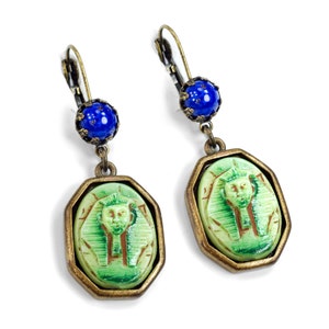 May include: A pair of gold tone earrings with green and blue glass beads. The green beads feature a raised image of an Egyptian pharaoh's head.