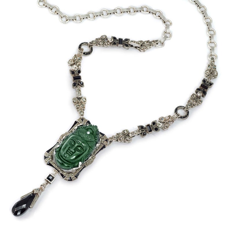 May include: A silver necklace with a green jade pendant carved with a face. The pendant is surrounded by black and white stones. The necklace is on a white background.