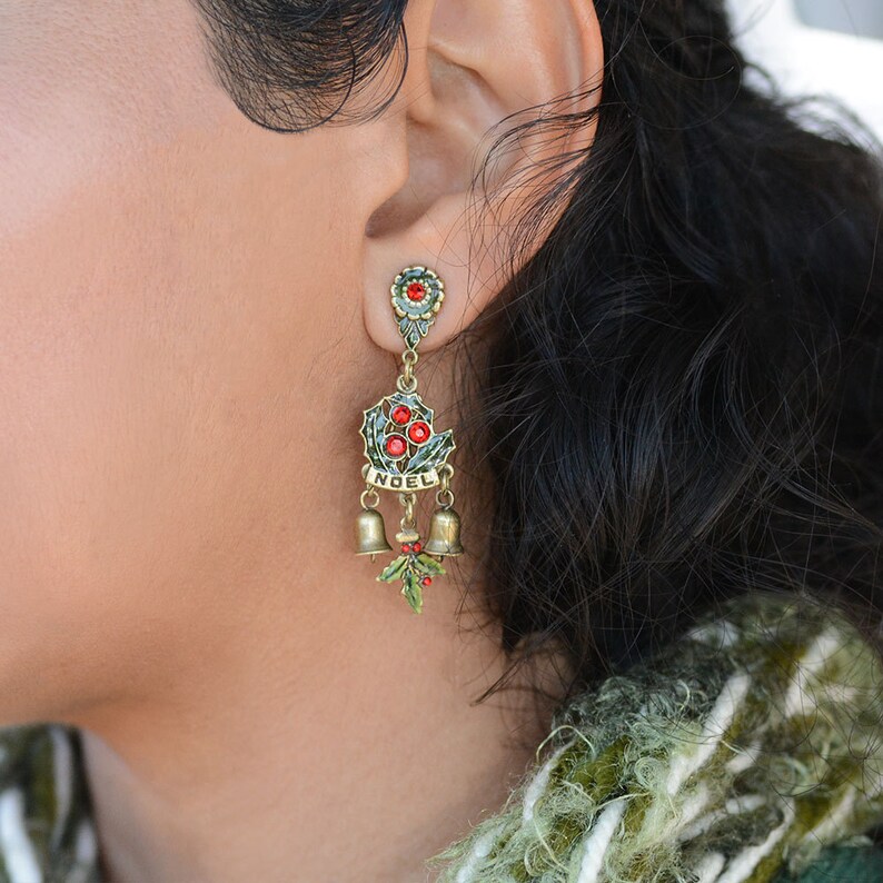 May include: Gold-tone dangle earrings with a green holly leaf design, red berries, and the word "NOEL" in gold lettering. The earrings feature three small gold bells.