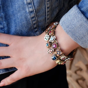 May include: A gold-tone bracelet with a chain link design and colorful, faceted gemstones. The bracelet features a variety of shapes and sizes of gemstones, including squares, circles, and ovals. The gemstones are set in gold-tone metal and are surrounded by intricate details.