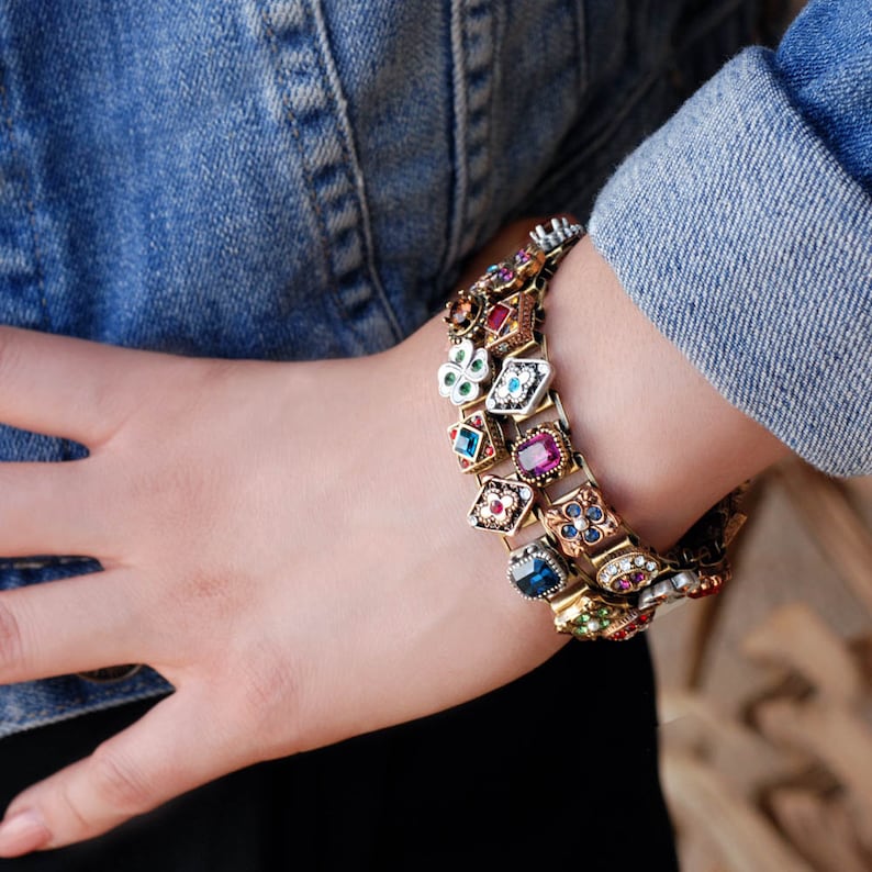 May include: A gold-tone bracelet with a chain link design and colorful, faceted gemstones. The bracelet features a variety of shapes and sizes of gemstones, including squares, circles, and ovals. The gemstones are set in gold-tone metal and are surrounded by intricate details.