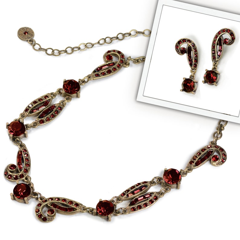 May include: A red crystal necklace and earrings set with a gold tone chain and ornate detailing. The necklace features a delicate chain with a pendant that has a paisley design. The earrings are drop earrings with a similar paisley design.