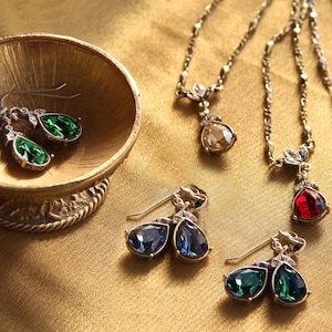May include: A set of three pairs of teardrop-shaped earrings with silver-tone settings and various coloured crystals. The earrings are displayed on a gold-coloured surface.