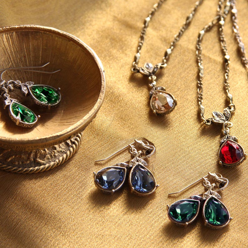 May include: A set of three pairs of teardrop-shaped earrings with silver-tone settings and various coloured crystals. The earrings are displayed on a gold-coloured surface.