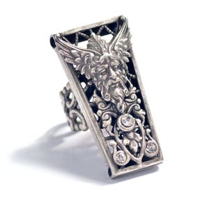 May include: A silver ring with a detailed design featuring a winged, bearded face and clear rhinestones.