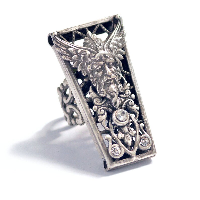 May include: A silver ring with a detailed design featuring a winged, bearded face and clear rhinestones.