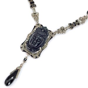 May include: A black and silver necklace with a carved black stone pendant featuring a Buddha face. The pendant is surrounded by a silver frame with black enamel accents. The necklace is adorned with black and clear crystals.