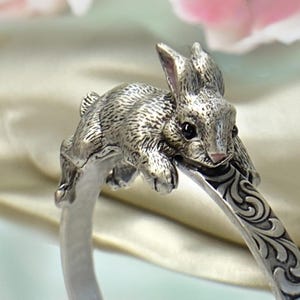 May include: A silver-toned ring featuring a detailed, textured bunny rabbit design. The rabbit is perched on the ring's band, which has an engraved floral pattern. The ring's design is intricate and detailed, with a focus on the rabbit's features.