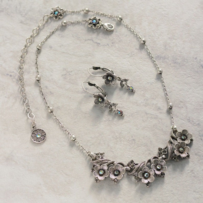Op de afbeelding: Een zilveren ketting en oorbellenset met een bloemenontwerp. De ketting heeft een delicate ketting met een hanger van verschillende met elkaar verbonden bloemen met iriserende stenen. De oorbellen zijn hangend met een enkele bloem en een iriserende steen.