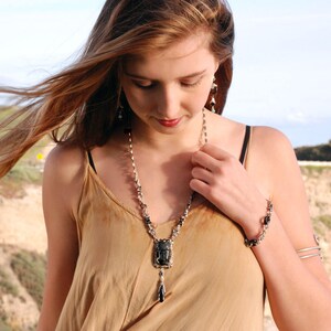 May include: A woman wearing a brown tank top with a silver chain necklace featuring a black stone pendant. The necklace has a long, dangling drop with a black stone at the end. She is also wearing a silver bracelet on her right wrist.