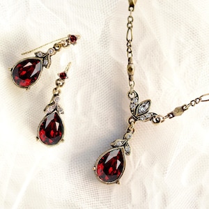Puede incluir: Un conjunto de joyas de estilo vintage que incluye un collar y pendientes a juego. El conjunto tiene un acabado dorado y está adornado con piedras preciosas rojas oscuras en forma de lágrima y pequeños cristales transparentes. Los pendientes son colgantes.