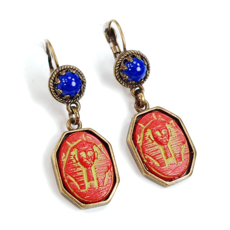 May include: A pair of red and blue dangle earrings with a gold tone finish. The earrings feature a red octagonal shaped pendant with a raised image of an Egyptian pharaoh. The top of the earrings feature a blue round bead.
